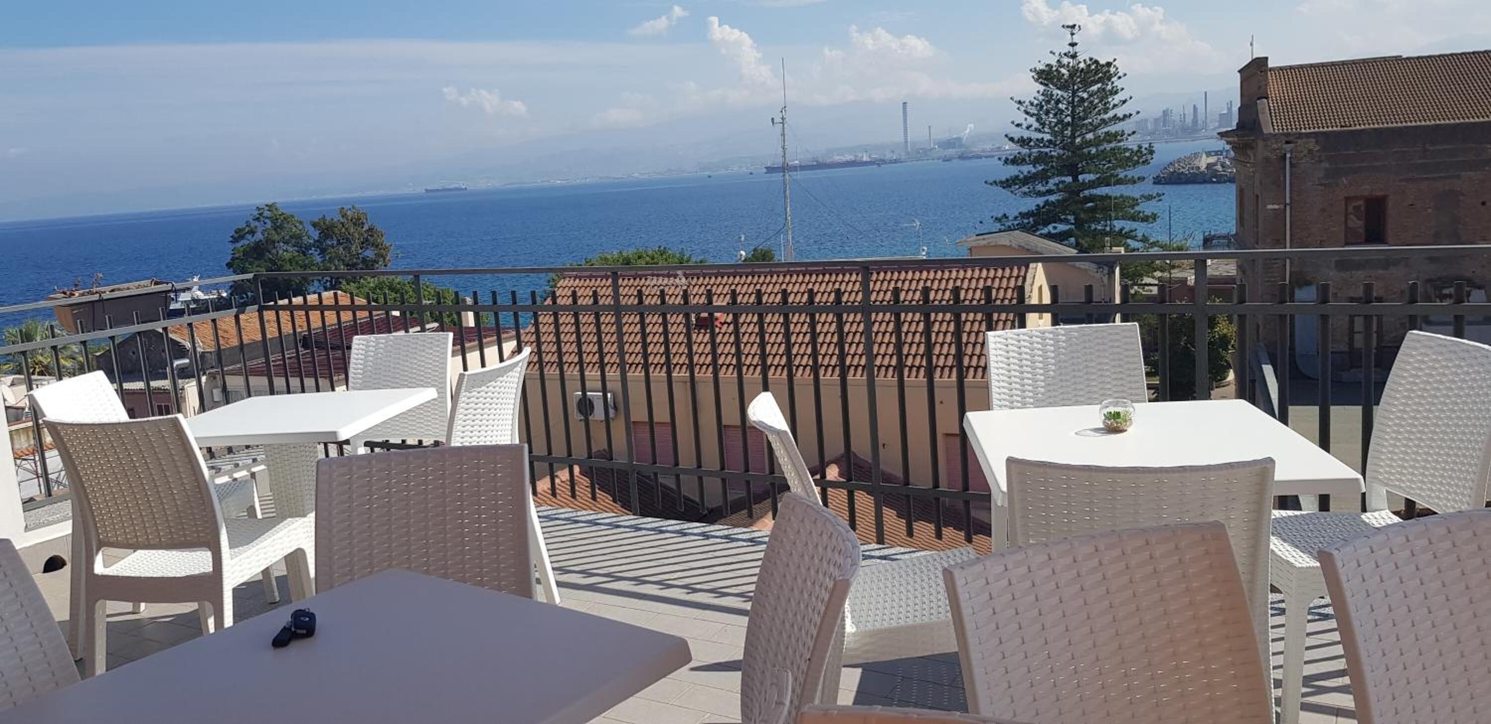 An outdoor terrace with white wicker furniture, a view of the sea, and a distant city skyline.