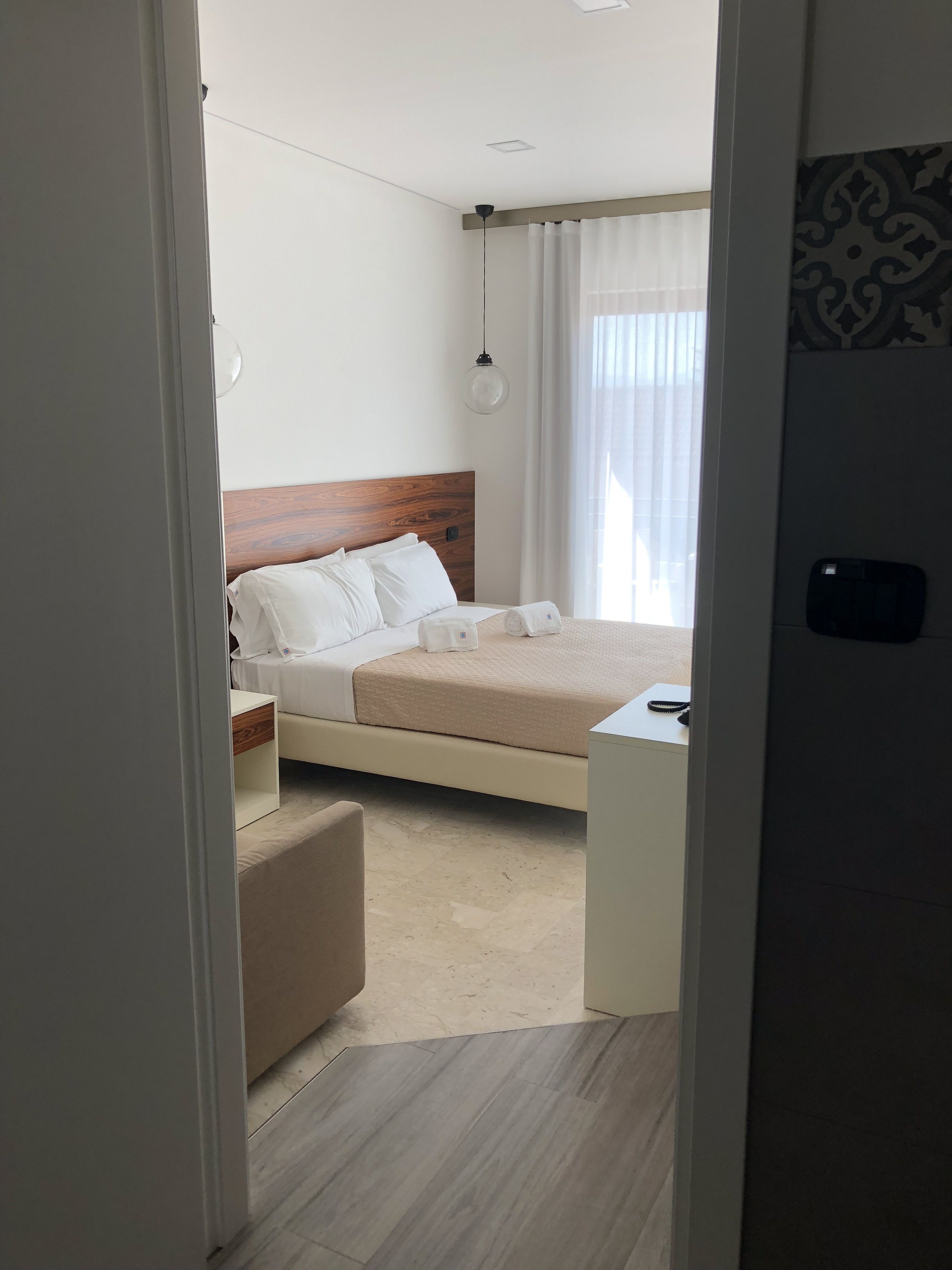 A modern hotel room with a wooden headboard, a bed with white linens, a bedside table, a small desk, and a window with curtains.
