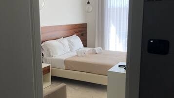 A modern hotel room with a wooden headboard, a bed with white linens, a bedside table, a small desk, and a window with curtains.