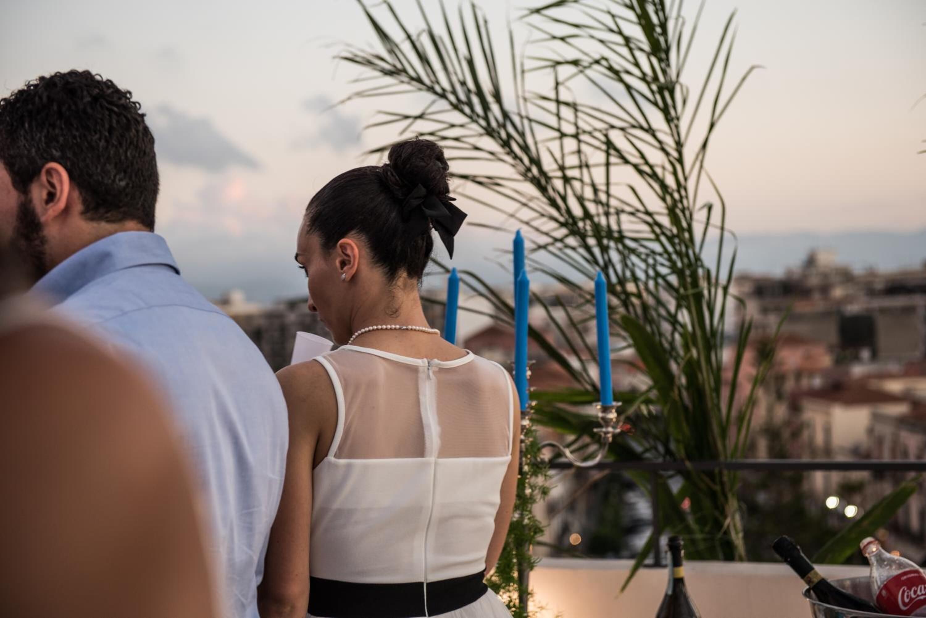 A couple standing on a balcony overlooking a cityscape, with a table featuring bottles and a candle.