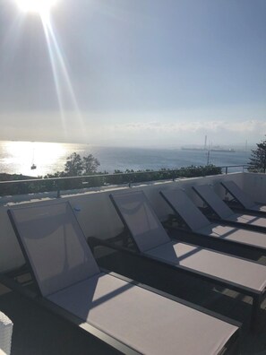 A rooftop terrace with sun loungers overlooking a body of water and distant industrial structures. - Milazzo INN AiMori (Milazzo)