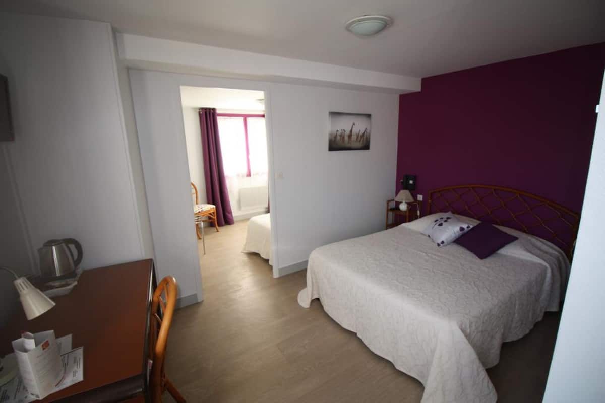 Una habitación de hotel con cama, escritorio con lámpara, silla, ventana con cortinas y un cuadro en la pared.