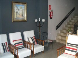 Un salón con tres sillones blancos, una mesita de madera, una lámpara de pie, un sillón con cojín estampado y una escalera. Se observa también un extintor en la pared.