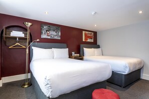 A hotel room with two beds, a red ottoman, a floor lamp, and artwork on the walls.