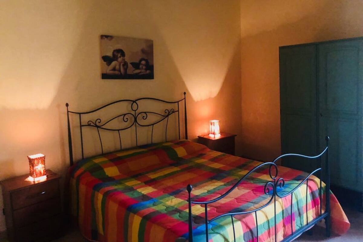 Un dormitorio con estructura de cama metálica, colcha colorida, mesita de noche con lámpara y armario verde.
