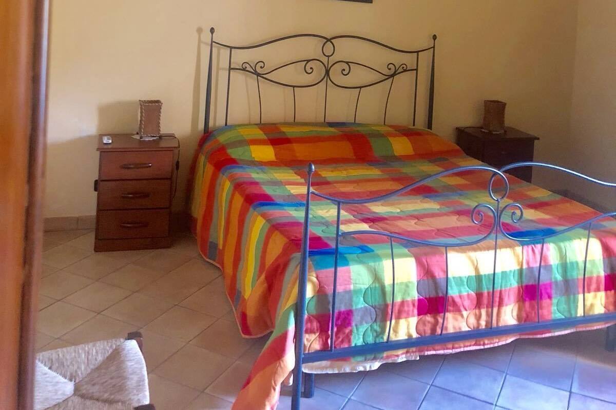 Un dormitorio con estructura de cama metálica, colcha colorida, una pequeña mesita de noche, lámpara de pared y un cuadro sobre la cama.