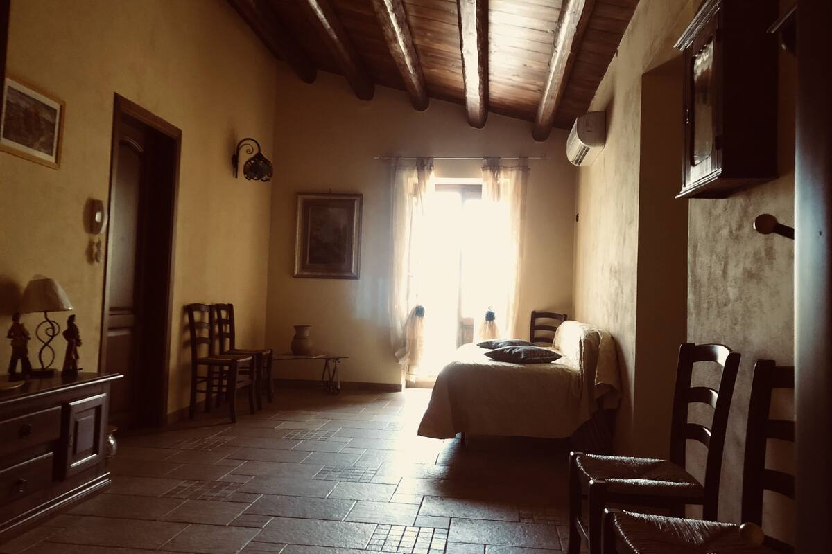 Una habitación con cama, sillas de madera, una mesita y un ventanal con cortinas.