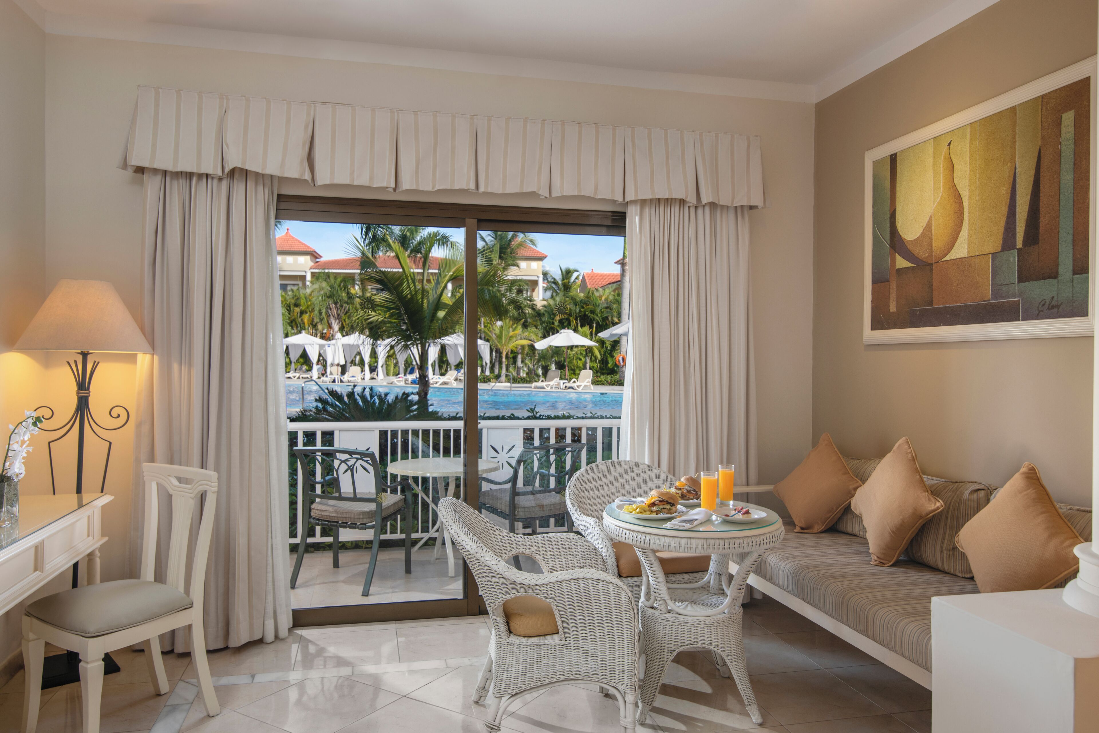 Habitación de hotel con balcón, muebles de mimbre, una mesa de comedor y un cuadro en la pared.