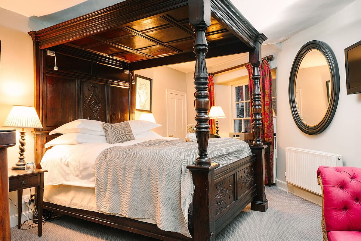 A four-poster bed with a herringbone-patterned bedspread, a wooden headboard, and a red upholstered chair.