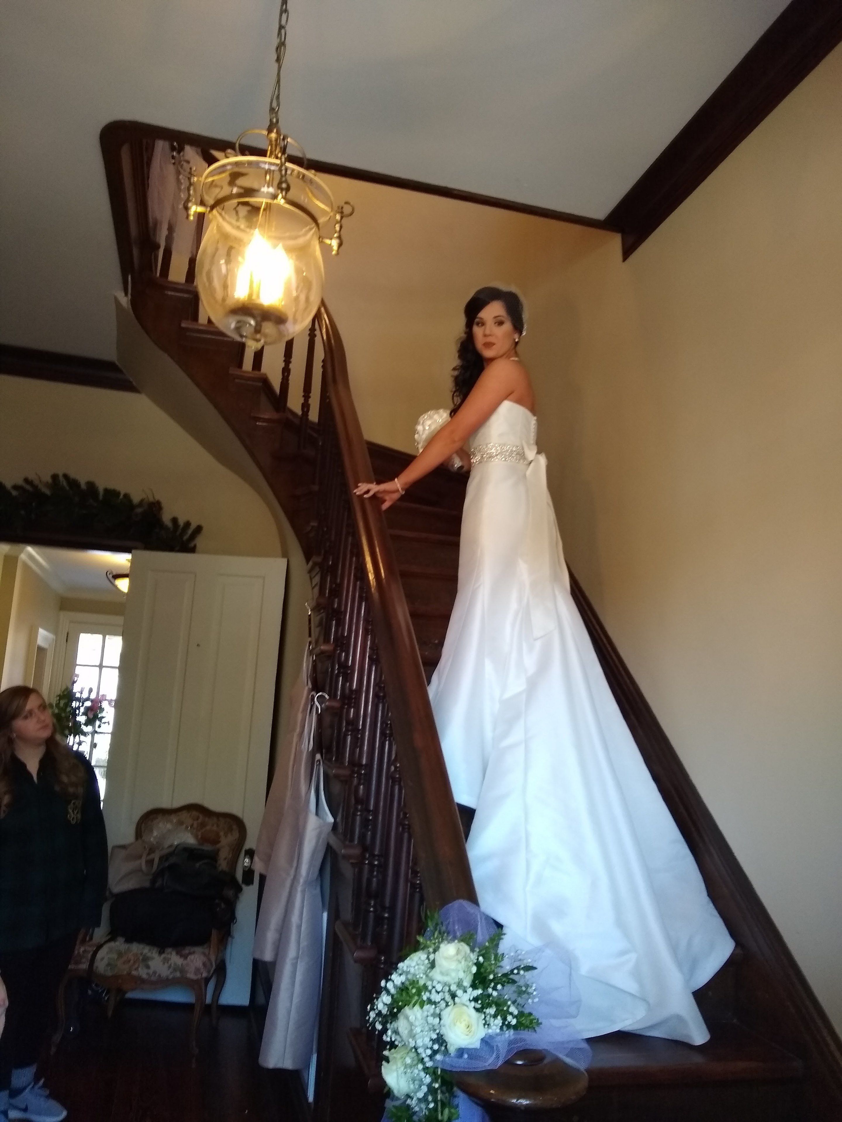 A staircase with a chandelier, a woman in a white dress, and a floral arrangement at the bottom.
