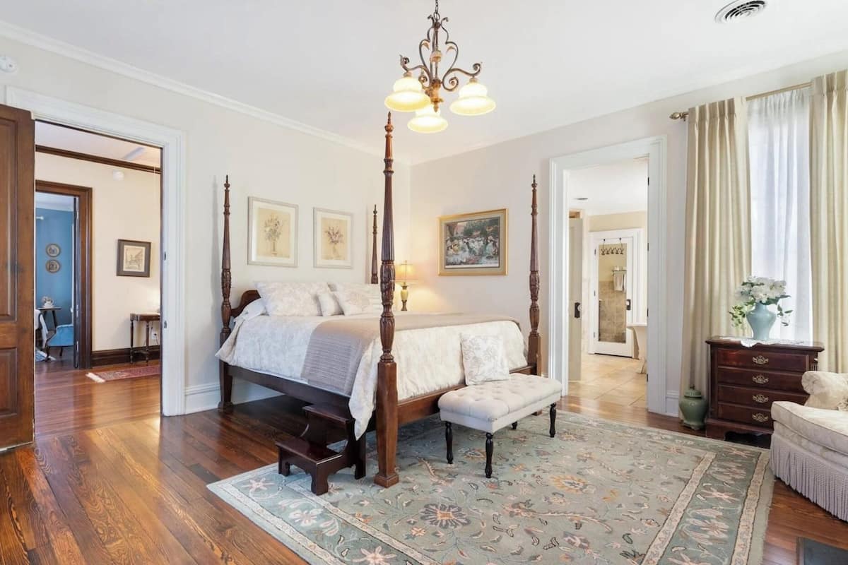 A spacious bedroom with a four-poster bed, a wooden floor, and a large window with curtains.