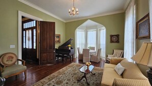 A living room with a grand piano, a sofa, a coffee table, and a chandelier. - The Wynne House Inn (Holly Springs)