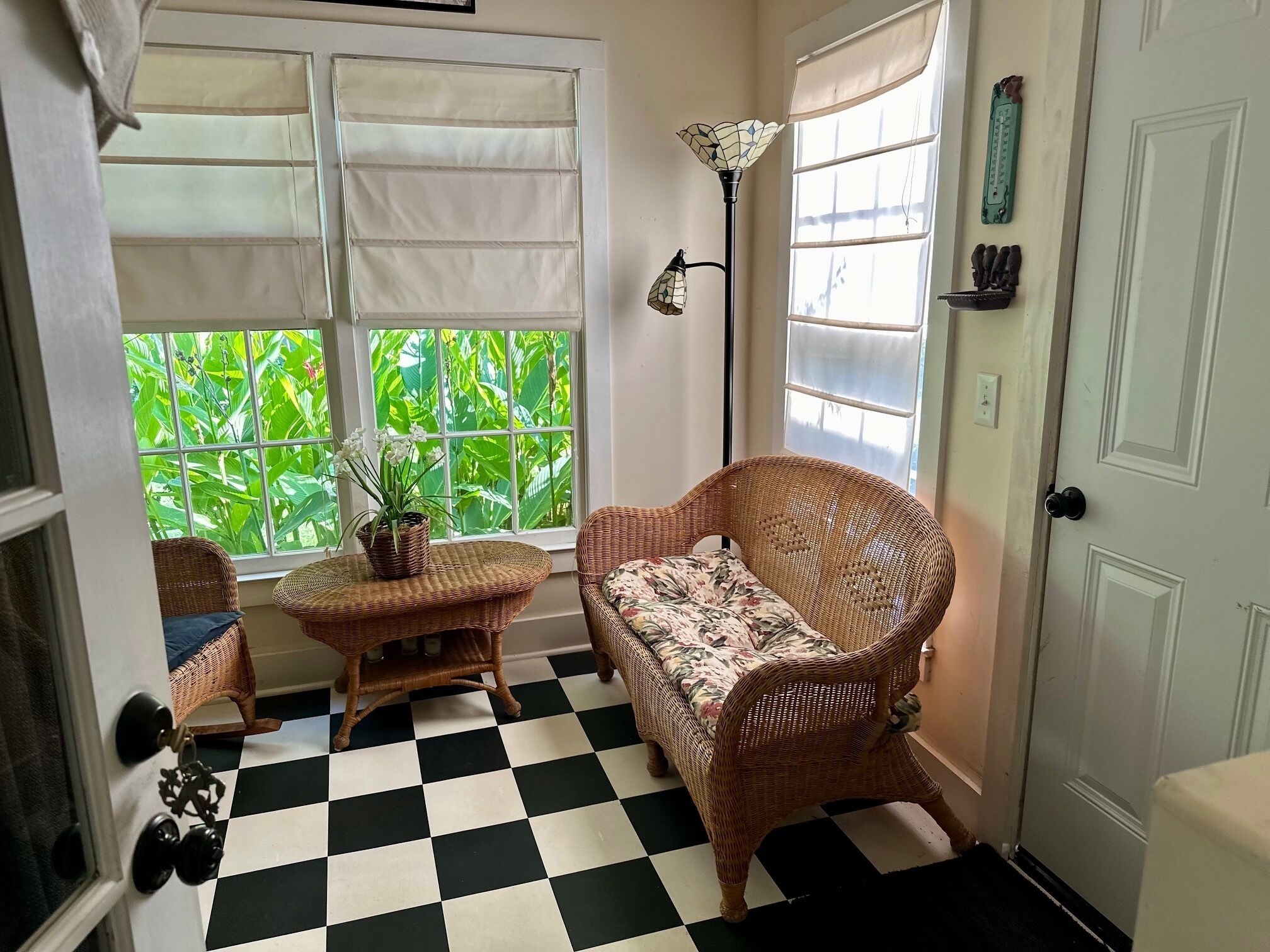 A room with a checkered floor, wicker furniture, a potted plant, and a window with a view of green foliage.