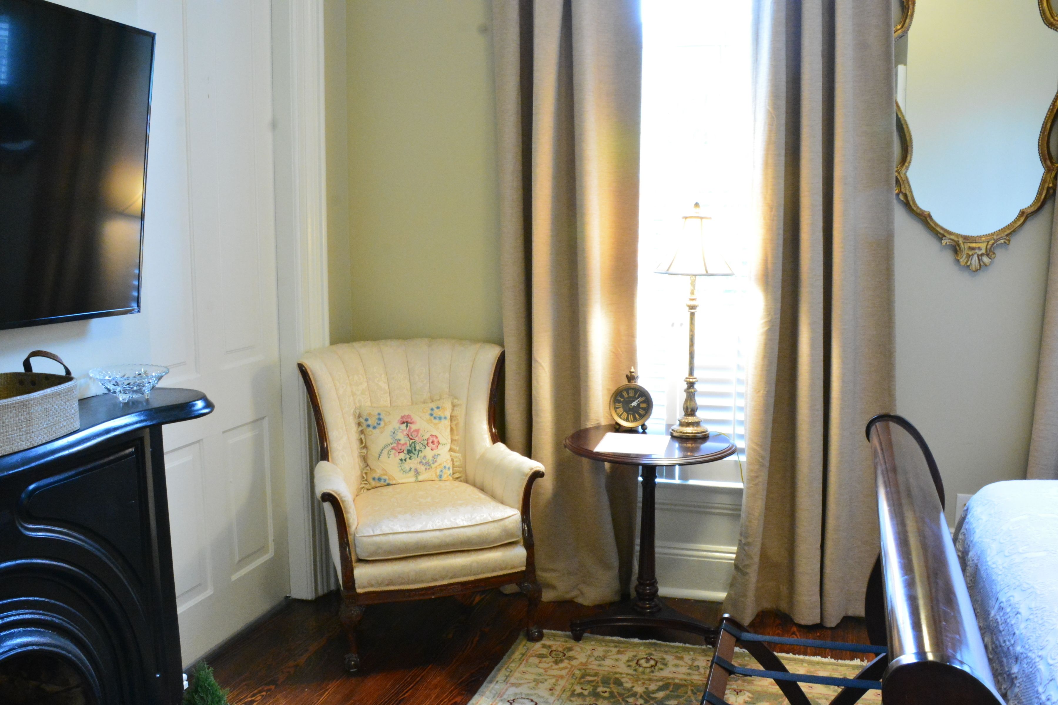 Une pièce avec un téléviseur fixé au mur, un fauteuil à motifs floraux, une petite table avec une lampe et un miroir doté d’un cadre orné.