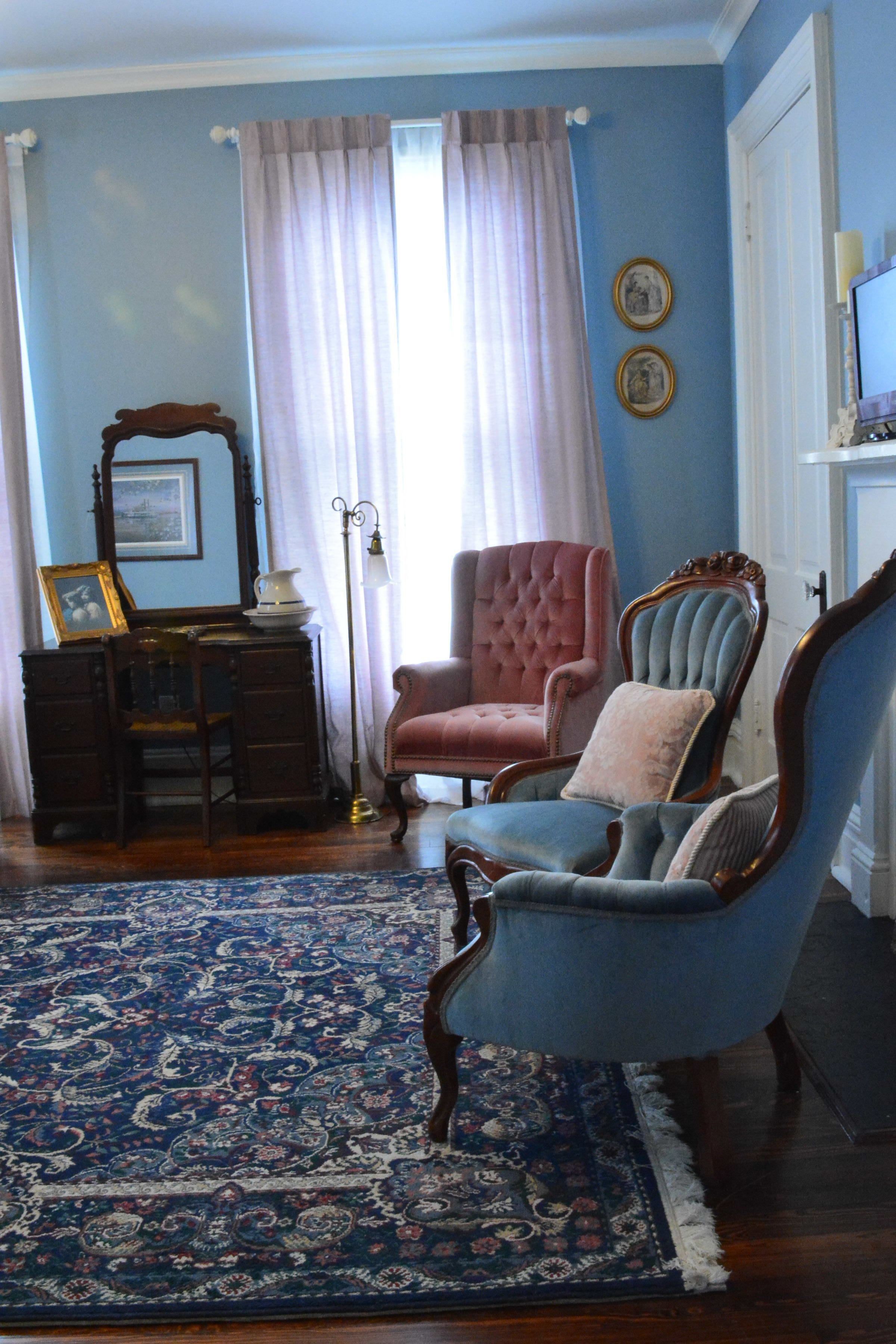 Una stanza con un muro blu, un comò in legno, una poltrona blu, una poltrona rosa, un tappeto decorato e uno specchio appeso al muro.