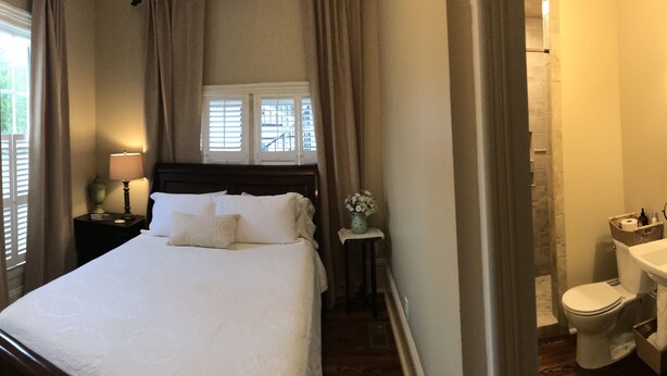A neatly arranged bedroom with a large bed, a bedside table with a lamp, a window with curtains, and a view of the outside. - The Wynne House Inn (Holly Springs)