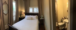 A neatly arranged bedroom with a large bed, a bedside table with a lamp, a window with curtains, and a view of the outside. - The Wynne House Inn (Holly Springs)