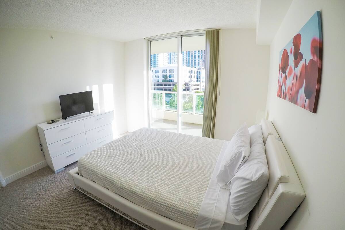 A bedroom with a bed, a TV on a dresser, a window with curtains, and a painting on the wall.