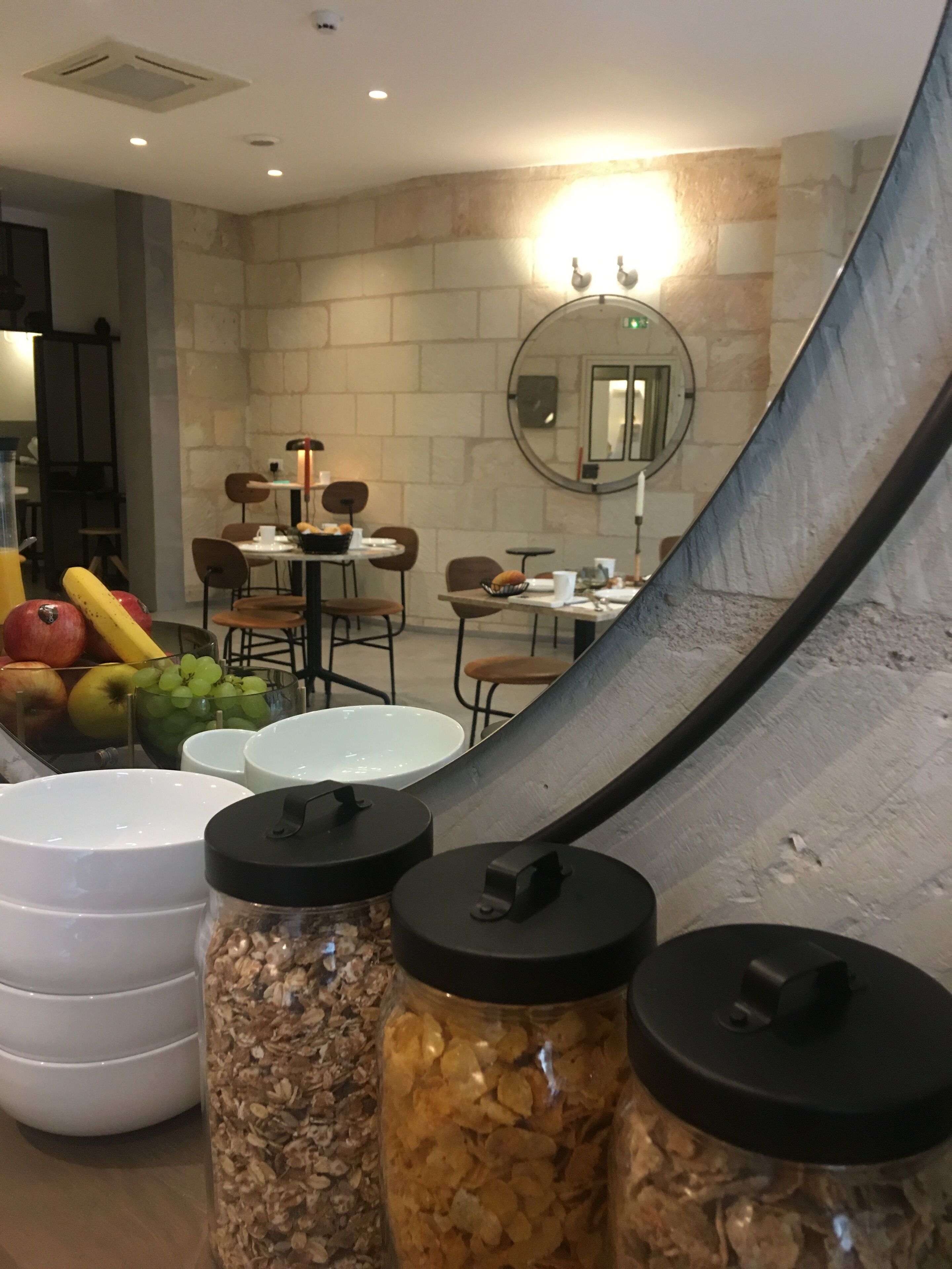 Un comedor con pared de piedra, mesas redondas, sillas y un espejo. Hay alimentos como frutas y frascos de cereales sobre la mesa.