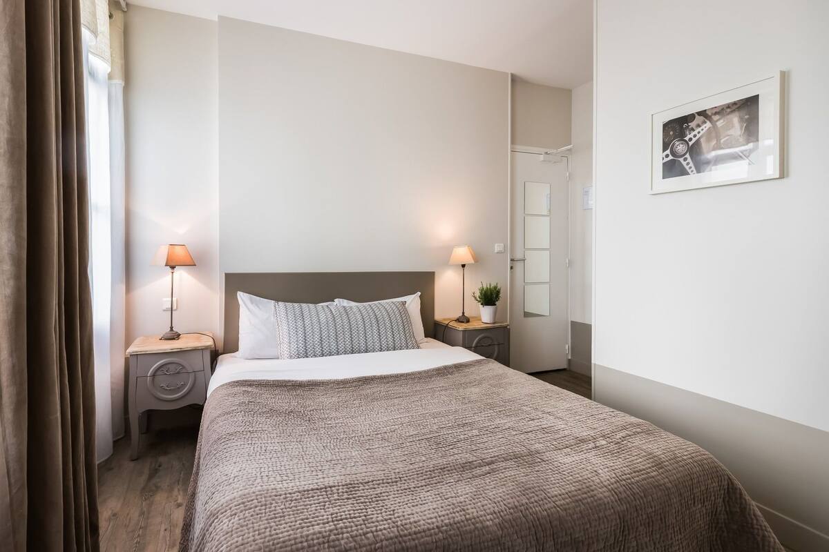 Une chambre d’hôtel bien rangée, avec un grand lit, deux lampes de chevet, une petite plante en pot et un tableau encadré au mur.
