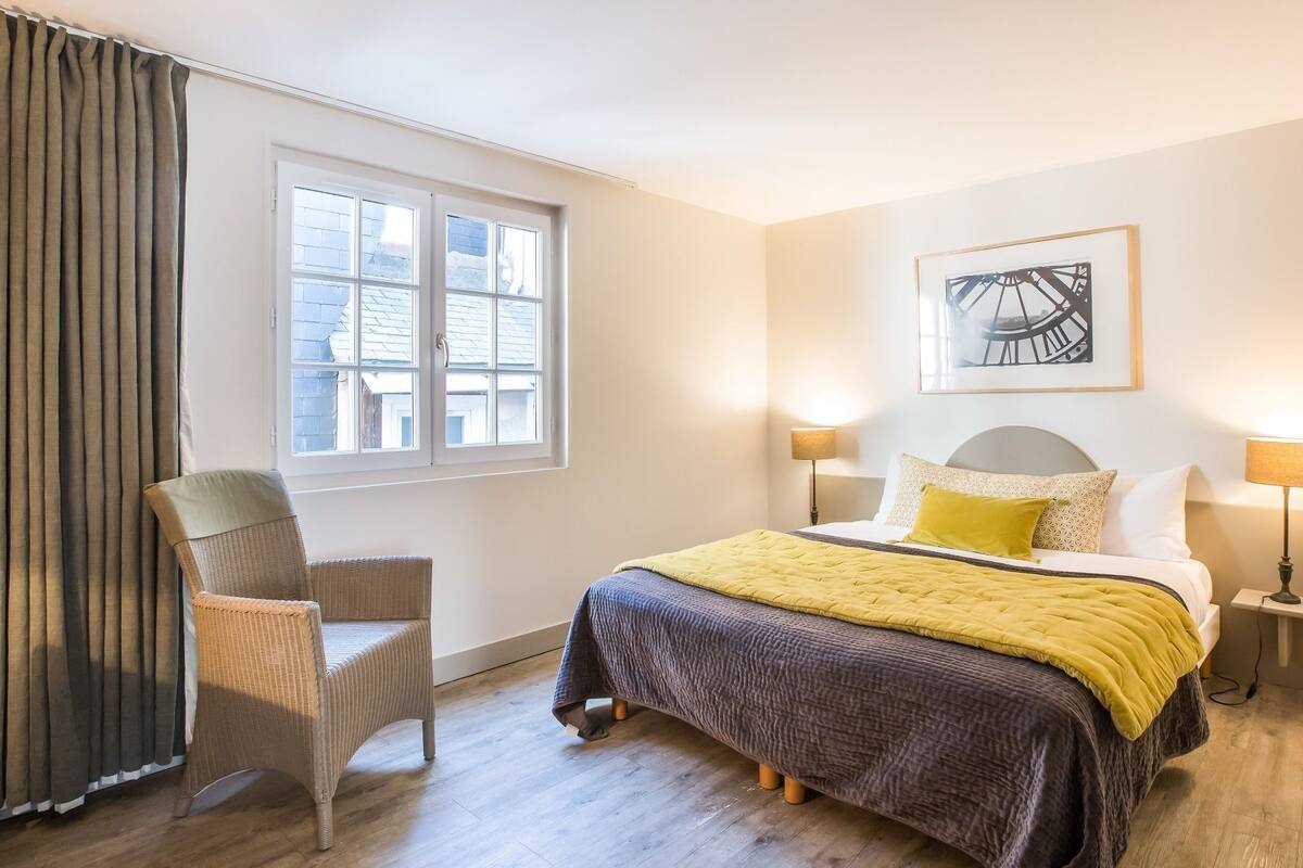 Une chambre d’hôtel comprenant un lit, deux lampes de chevet, une chaise, une fenêtre avec des rideaux et une œuvre d’art accrochée au mur.