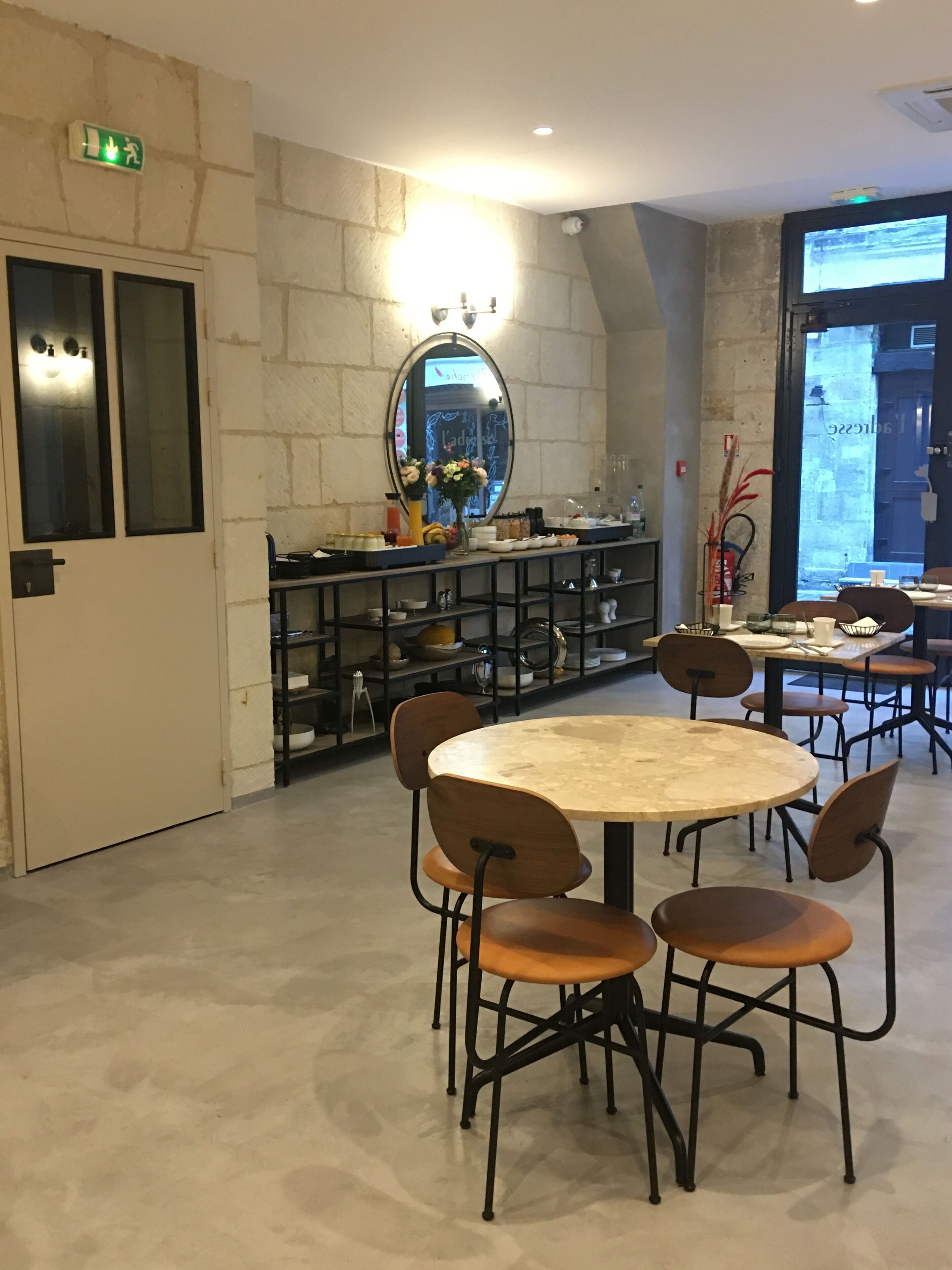 A dining area with a round table and chairs, a mirror, shelves with dishes, and a view through a large window.