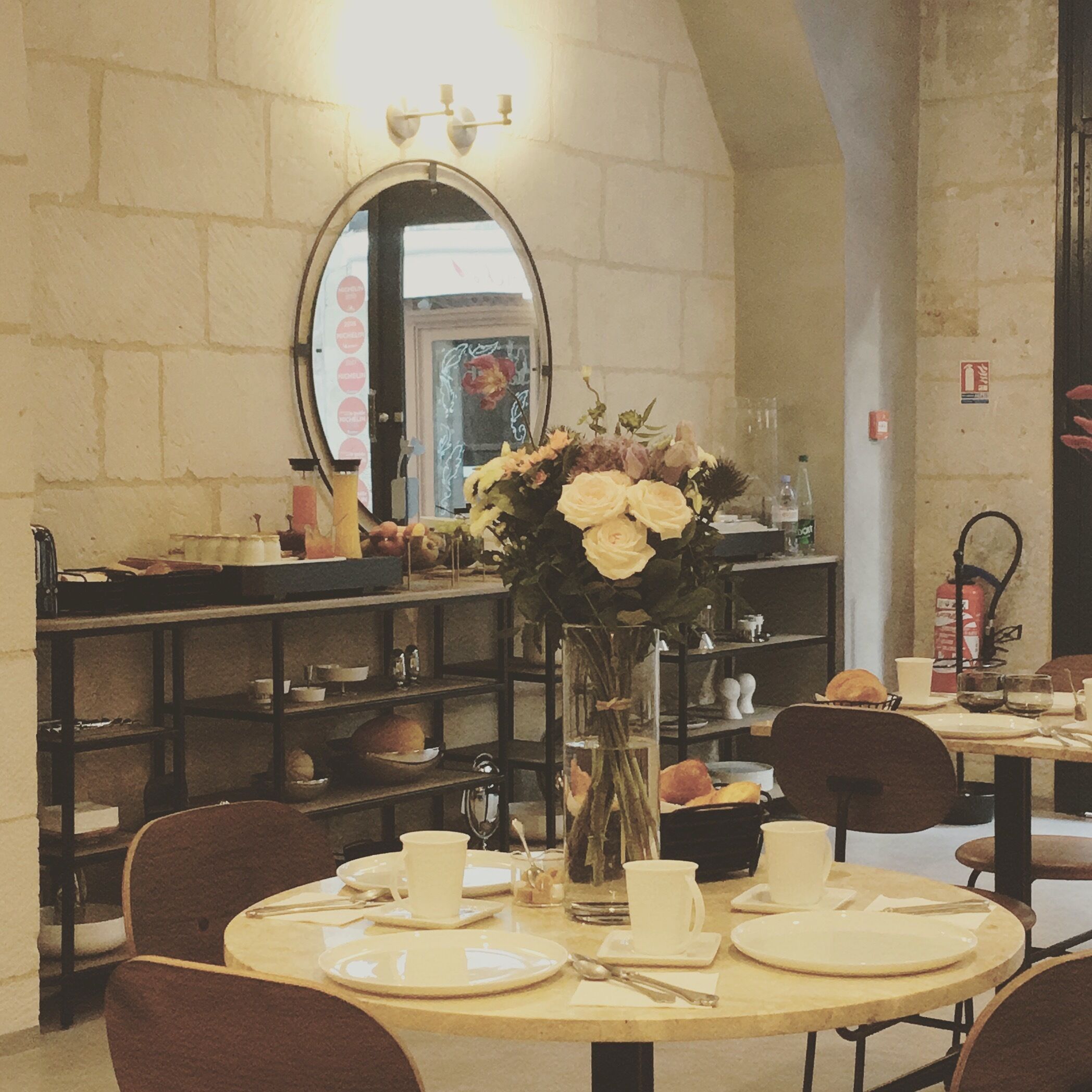 A dining area with round tables, chairs, and a vase with flowers. There is a mirror on the wall and shelves with various items.