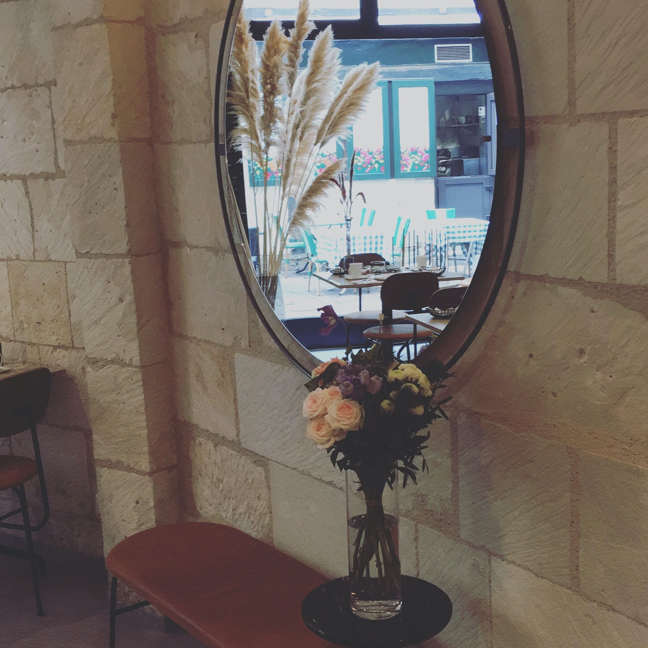 A mirror reflecting a room with a stone wall, a bench, and a vase with flowers.