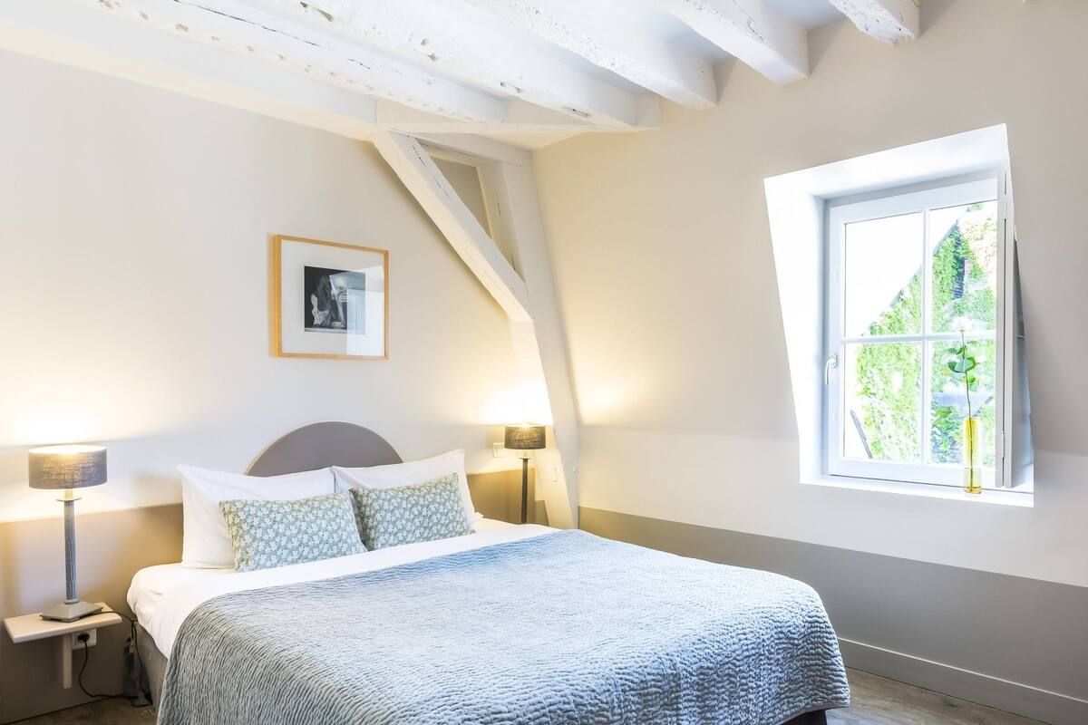 Une chambre à coucher avec un lit, deux lampes de chevet, un tableau encadré au mur et une fenêtre donnant sur un paysage verdoyant.