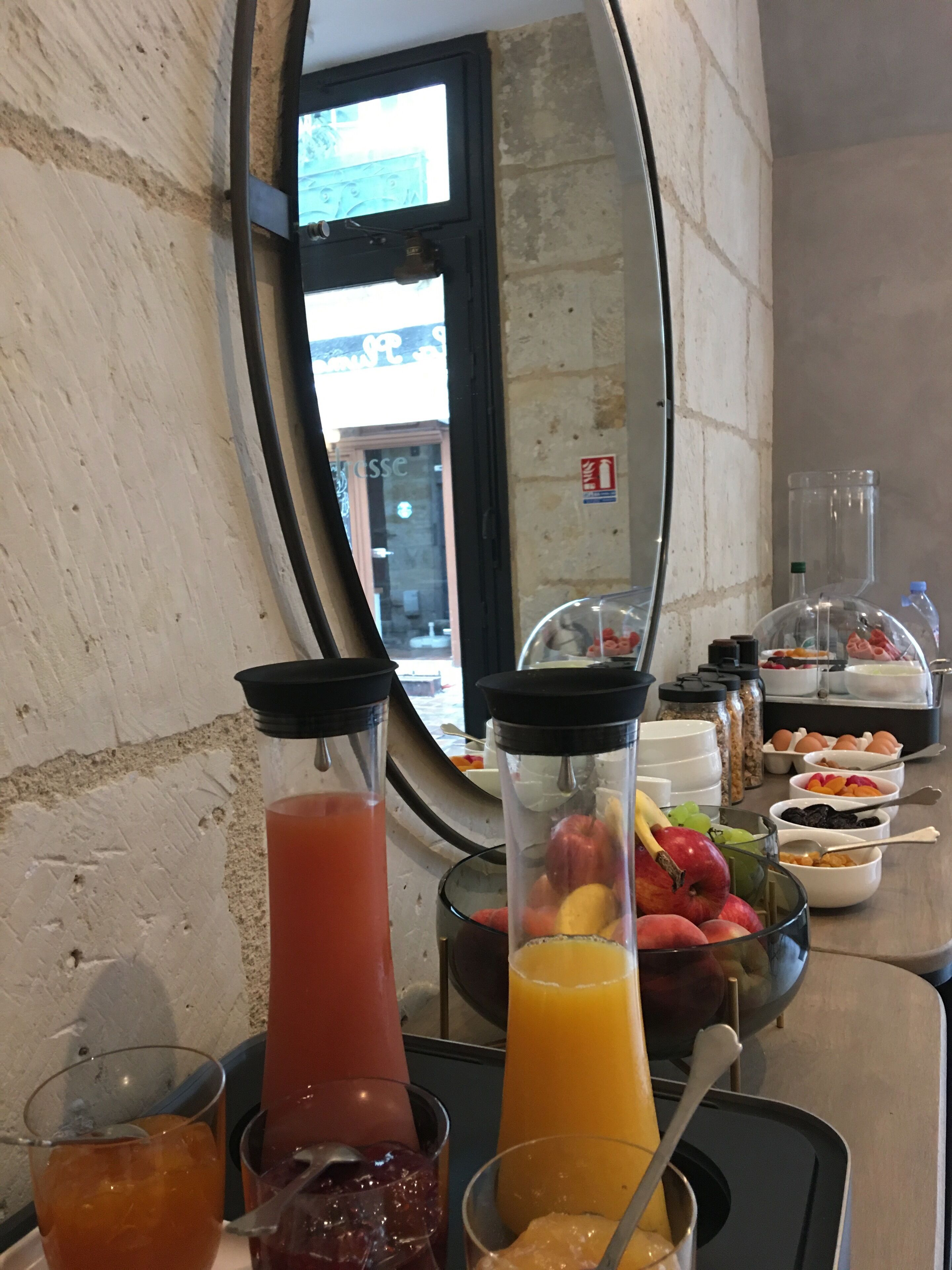 A breakfast buffet with various fruits, juices, and a mirror reflecting the room.