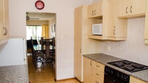 A compact kitchen with wooden cabinets, a microwave, a stove, and a dining area with chairs and a table. - Goulden Street Guest House (East London)