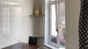 A dining area with a wooden table and chairs, a hanging light fixture, a window with a view of flowers, and a patterned curtain. - Exclusive Flats In Brussels - Olives (Brussels)