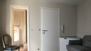 A room with a grey sofa, a black chair, a white door, and a small kitchenette area. - Exclusive Flats In Brussels - Olives (Brussels)
