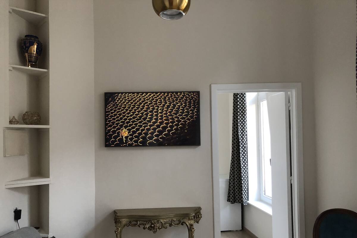 A room with a white wall, a black and gold artwork, a patterned curtain, a small table, and a chair.