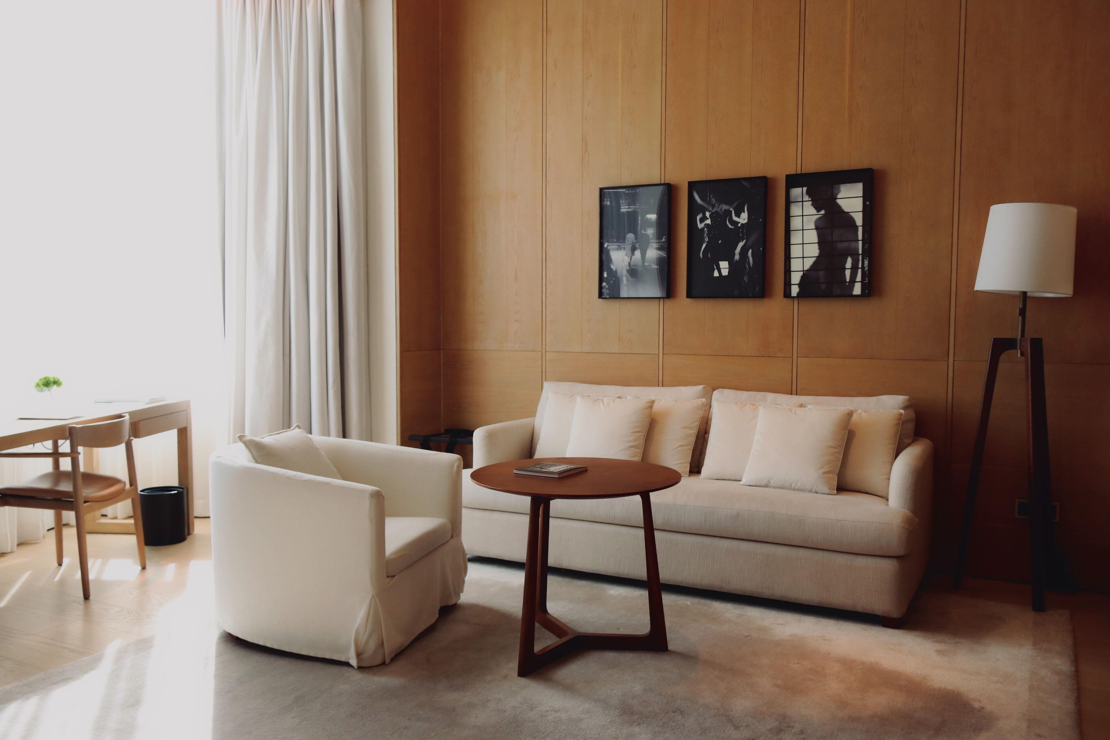 Una sala de estar moderna con un sofá, un sillón y una mesa de centro redonda. Hay tres cuadros enmarcados en la pared, una lámpara de pie y una ventana con cortinas.