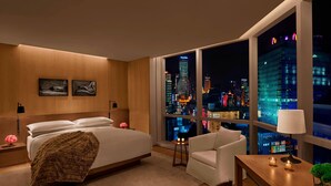A modern hotel room with a large bed, a sitting area with a table and chair, a nightstand, and a view of the city skyline at night. - The Shanghai EDITION (Shanghai)