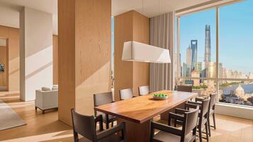 A modern hotel room with a wooden dining table, six chairs, a large window with a city view, and a white lamp.