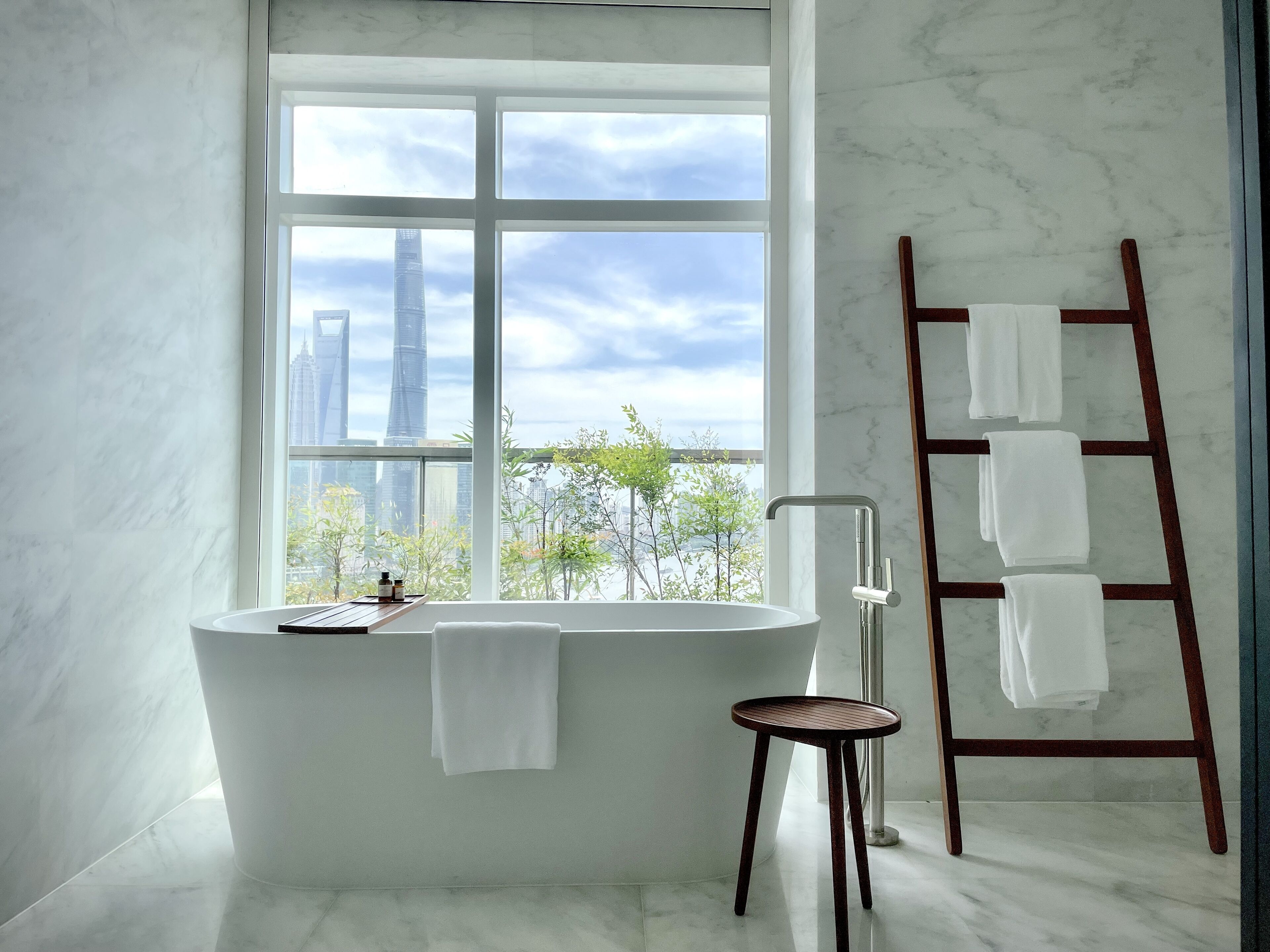Une salle de bain avec une baignoire sur pieds, un tabouret en bois, un porte-serviettes et une vue sur le paysage urbain, visible à travers une grande fenêtre.