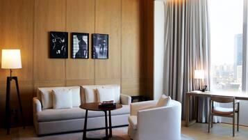 Una habitación de hotel moderna con sofá, sillón y escritorio. Hay tres cuadros en la pared, una lámpara y vistas a la ciudad a través de amplios ventanales.