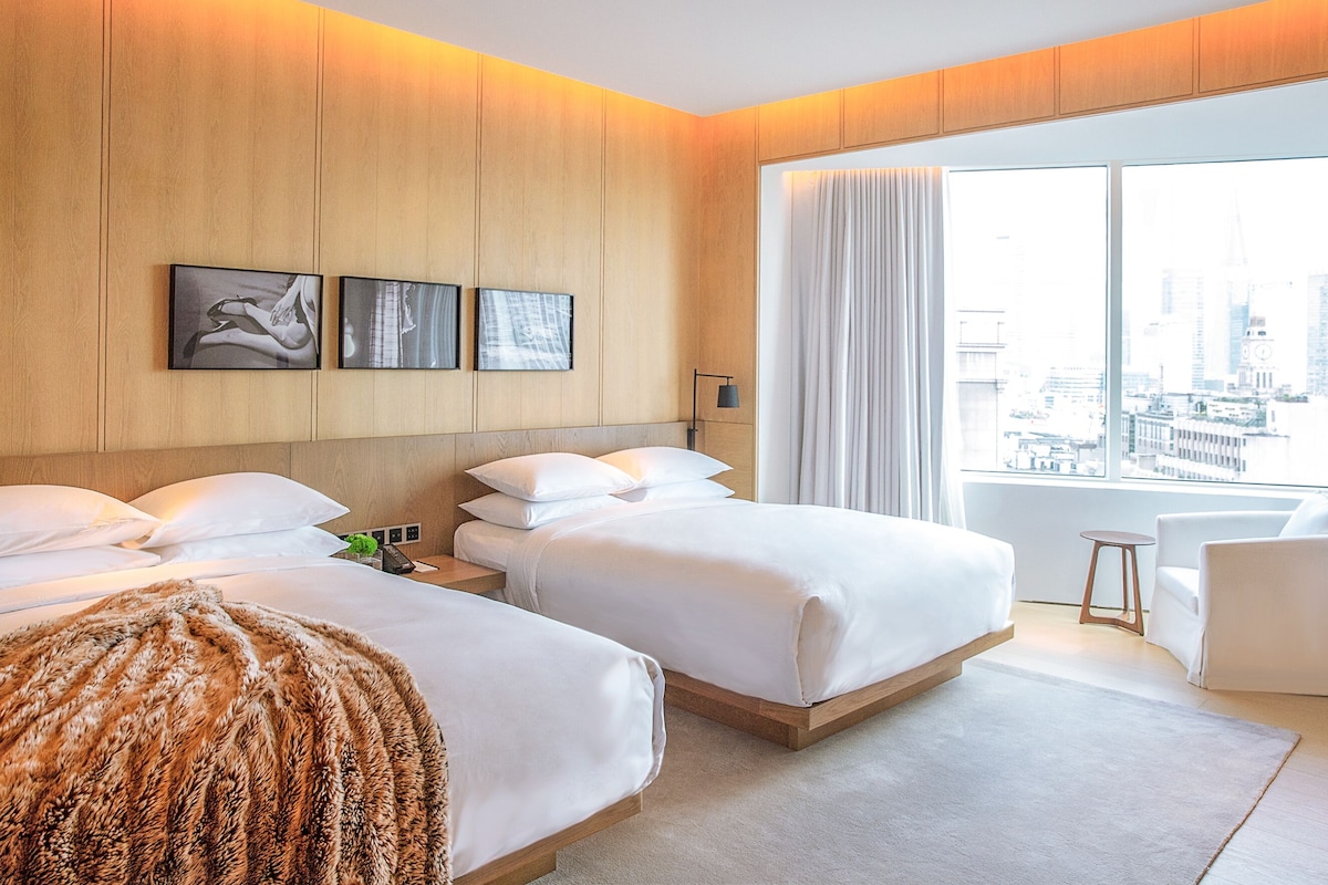 Hotellihuone, jossa on kaksi sänkyä, suuri ikkuna, josta on näkymä kaupunkiin, ja taideteoksia seinällä.