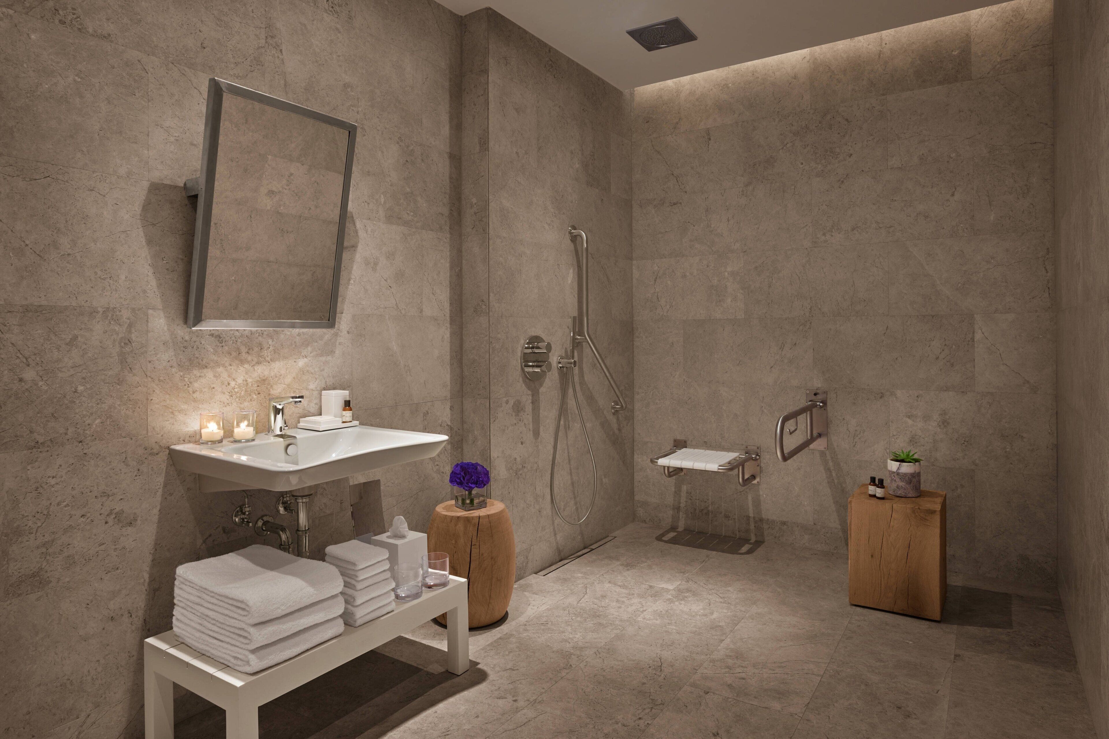 A modern bathroom with a large mirror, a sink, a shower, and a toilet. There are towels stacked on a bench, a small wooden stool with a plant, and a candle on the sink.