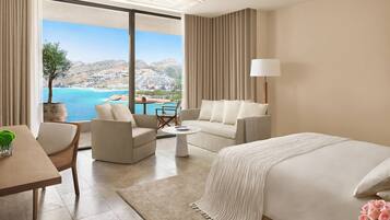 Habitación de hotel con una cama grande, dos sofás, una mesa de comedor y vistas al mar y a las colinas.