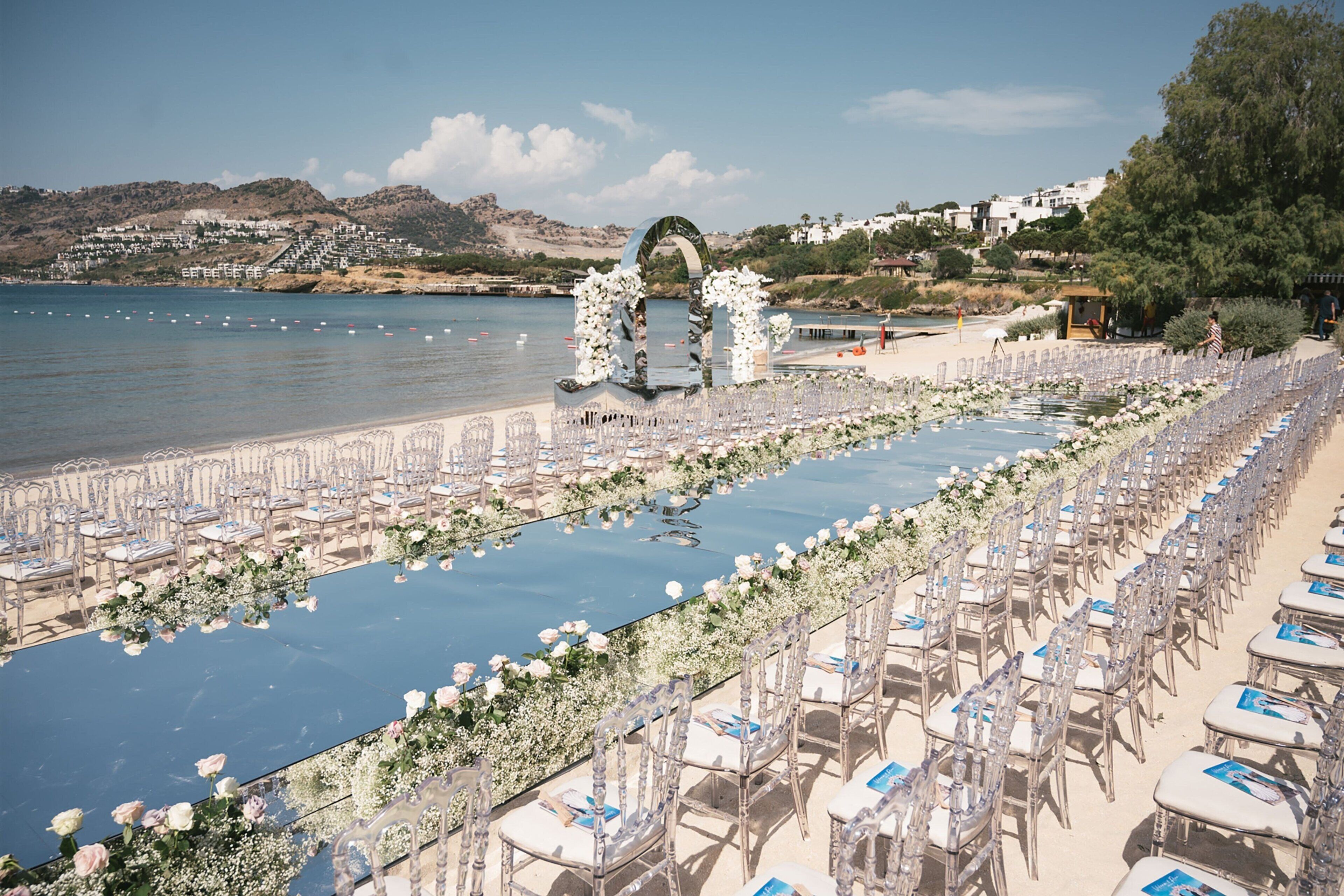 Une cérémonie de mariage organisée sur la plage, avec des rangées de chaises et une longue table décorée de fleurs, le tout offrant une vue sur la mer.