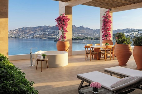 A terrace with a view of the sea, a table with chairs, a sofa, a bathtub, and large potted plants.