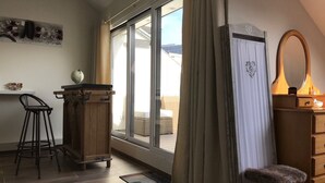 Ein Zimmer mit einer großen Glasschiebetür, die zu einem Außenbereich führt, einem Holztisch mit Stuhl, einer Kommode mit Spiegel und einem kleinen Hocker.