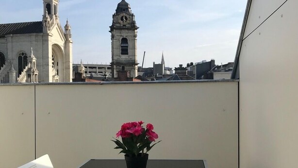 Ein Balkon mit Tisch und Stühlen, einem Blumentopf und Blick auf ein historisches Gebäude mit einem Glockenturm.