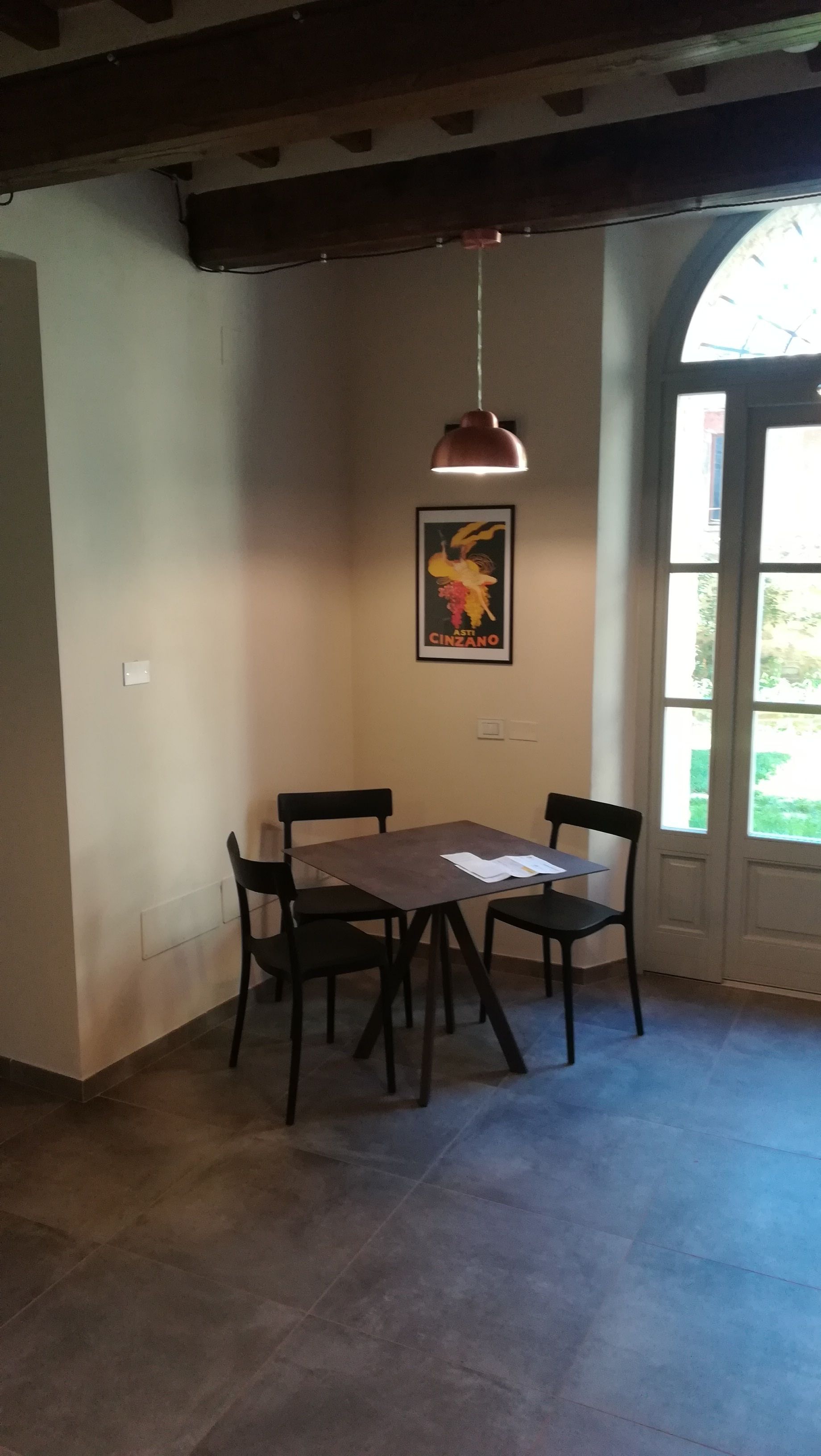 Zona pranzo con tavolo e quattro sedie, una lampada a sospensione, un quadro appeso e una finestra vista verde.