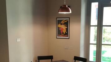 Zona pranzo con tavolo e quattro sedie, una lampada a sospensione, un quadro appeso e una finestra vista verde.