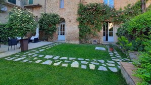 Une cour avec un chemin de pierre, une pelouse verdoyante et un bâtiment aux murs de pierre et aux nombreuses fenêtres.