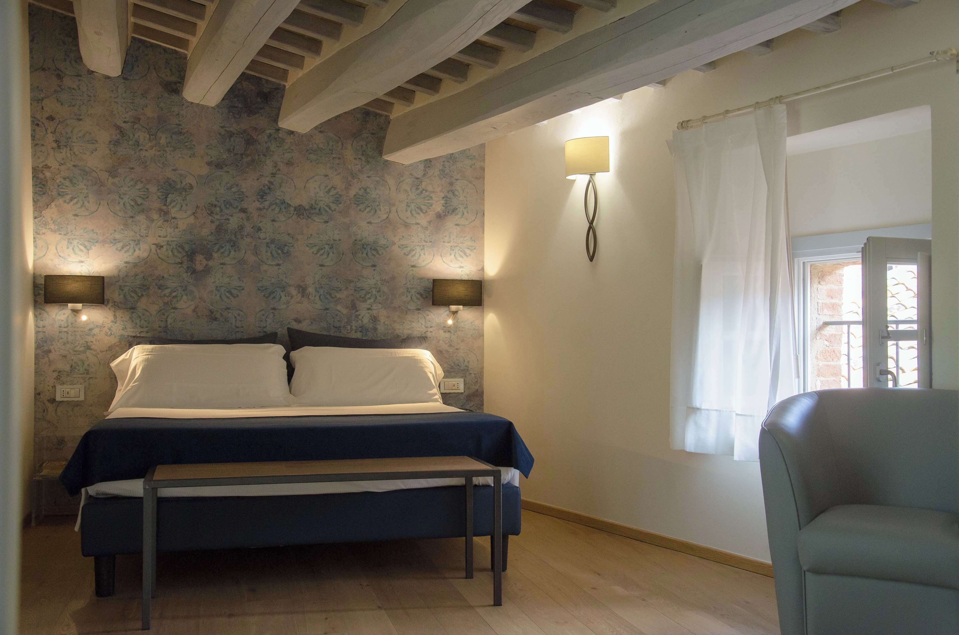 Un dormitorio con cama, lámparas de noche, un banco, una ventana con cortinas y una lámpara de pared.